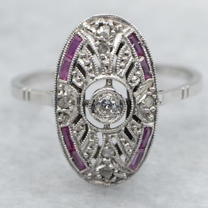 Può includere: Anello ovale in argento con un design a filigrana dettagliato. Presenta un diamante centrale, diamanti più piccoli e rubini rettangolari. Un gioiello in stile vintage, adatto per occasioni speciali.