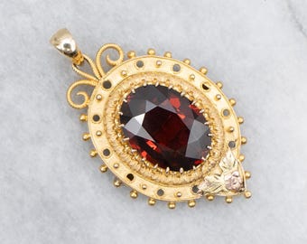 Könnte beinhalten: Goldener Anhänger mit einem großen, oval geschliffenen, tiefroten Edelstein. Der Anhänger hat eine verzierte Goldeinfassung mit dekorativem Rand und kleinem floralen Akzent. Der Bügel ist eine einfache Schlaufe.