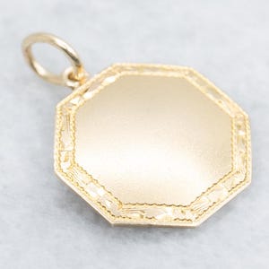May include: A gold-coloured pendant with an octagonal shape. The pendant has a smooth, blank centre and a decorative border. A small loop is attached at the top for a chain. The pendant is likely made of gold or a gold-toned metal.