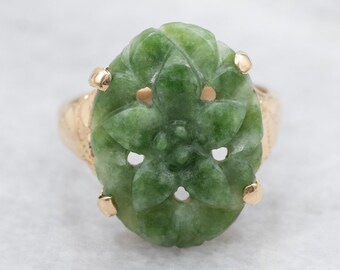 May include: A gold ring featuring a carved green jade stone. The jade is oval-shaped with a floral design and is set in a gold band. The ring has a vintage aesthetic.