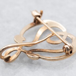 May include: A gold-toned brooch in the shape of a stylized heart with a delicate, intricate design. The brooch has a pin and catch closure.