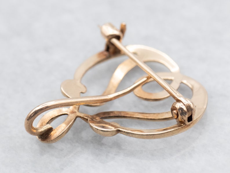 May include: A gold-toned brooch in the shape of a stylized heart with a delicate, intricate design. The brooch has a pin and catch closure.
