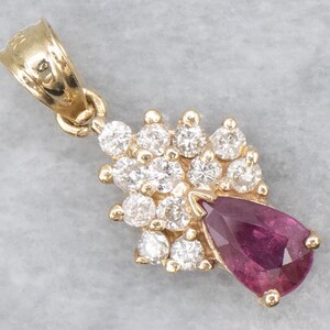 May include: A gold pendant featuring a pear-shaped deep pink gemstone at the base, surrounded by a cluster of twelve round clear stones. The pendant includes a polished gold bail at the top for attachment to a chain, showcasing a classic design.