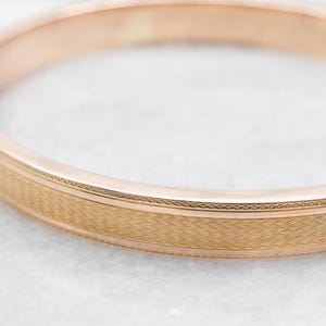 May include: A close-up shot of a gold-colored bangle bracelet. The bracelet has a textured band with a repeating wave pattern. The edges of the band are bordered by a thin, decorative line. The bracelet is set against a blurred, light-colored background.