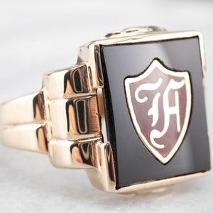 Puede incluir: Un anillo de oro con una piedra de ónix negro engastada en forma cuadrada. La piedra presenta un escudo con las iniciales "FA" en blanco.