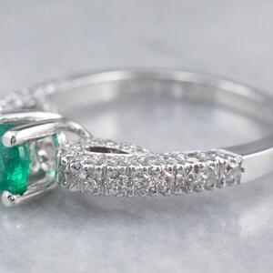 May include: A silver ring featuring a large emerald gemstone set within a halo of smaller diamonds. The ring has a delicate and intricate design.