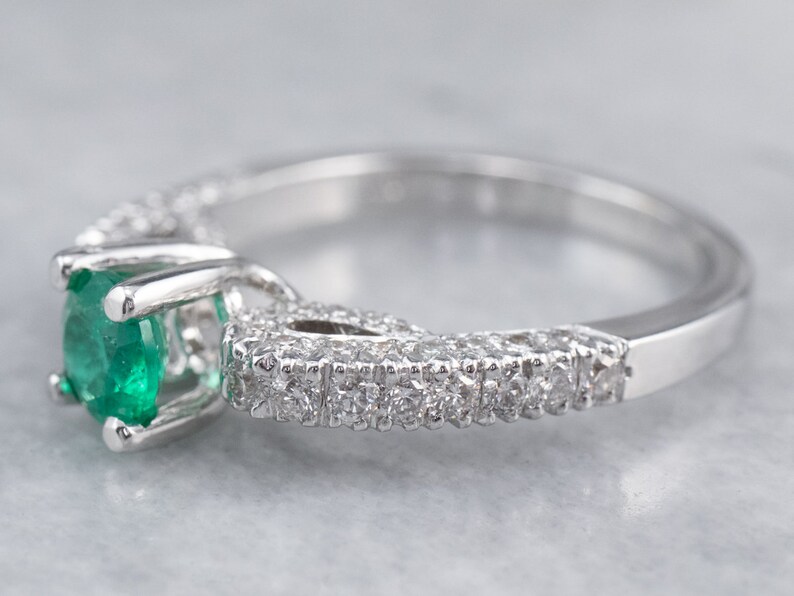 May include: A silver ring featuring a large emerald gemstone set within a halo of smaller diamonds. The ring has a delicate and intricate design.