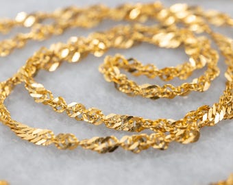 May include: Close-up of a gold-colored necklace with a twisted rope design. The chain is made of small, flat, interconnected links that create a textured surface. The necklace is displayed on a light gray surface.