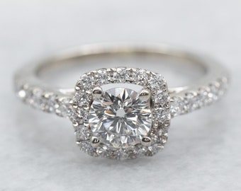 May include: A silver engagement ring with a large, round, center diamond surrounded by a double halo of smaller diamonds. The band is also set with smaller diamonds.