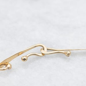 May include: Close-up of a gold-colored necklace with a unique design. The necklace features curved, elongated links with small, spherical accents. The links are connected in an intricate pattern, creating an elegant and eye-catching piece of jewelry.