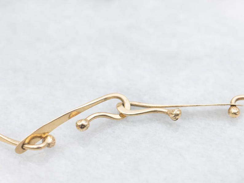 May include: Close-up of a gold-colored necklace with a unique design. The necklace features curved, elongated links with small, spherical accents. The links are connected in an intricate pattern, creating an elegant and eye-catching piece of jewelry.