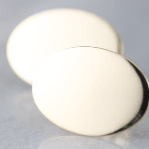 Puede incluir: Dos objetos dorados de forma ovalada, uno ligeramente más grande que el otro, están colocados cerca uno del otro. Los objetos tienen una superficie lisa y reflectante, con un borde oscuro. El fondo es de un color suave y neutro.