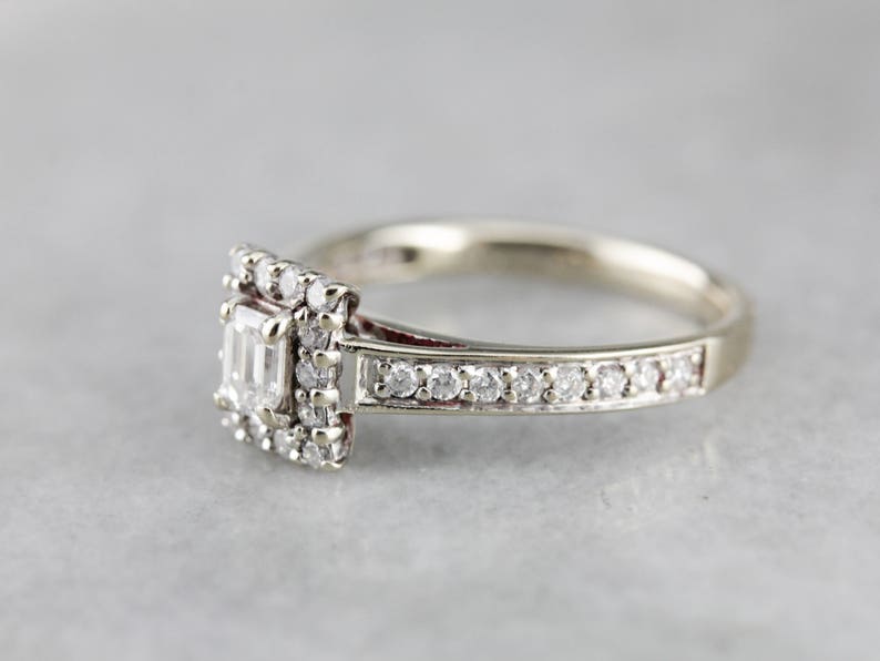 Puede incluir: Un anillo de compromiso de plata con un diamante central de forma cuadrada rodeado de diamantes m&aacute;s peque&ntilde;os. La banda tambi&eacute;n est&aacute; engastada con peque&ntilde;os diamantes.