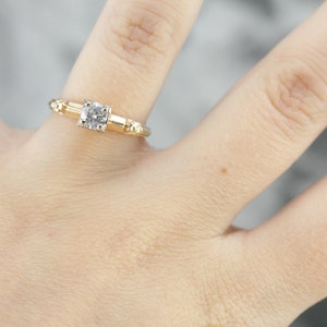 May include: A gold engagement ring with a round, clear diamond set in a simple band. The band is accented with small, round beads.