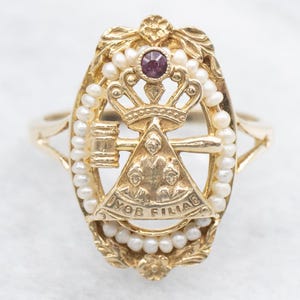 Op de afbeelding: Gouden ring met een paarse steen en een ontwerp van een kroon, een hamer en een driehoek met de woorden "MOB FILIAE" eromheen. De ring is omgeven door een rij parels.