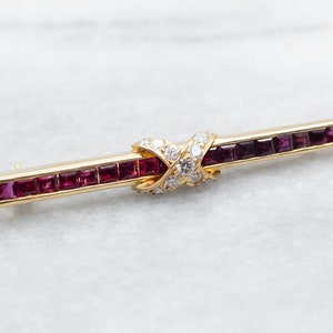 Può includere: Una spilla a barra d'oro con un designo di rubini e diamanti. La spilla presenta un nodo di diamanti al centro ed è bordata da pietre di rubino.