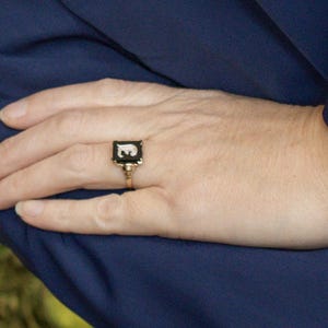 May include: A gold-toned ring with a black rectangular face featuring a white letter "E". The ring is set against a dark blue fabric background. The ring is a classic piece of jewelry.