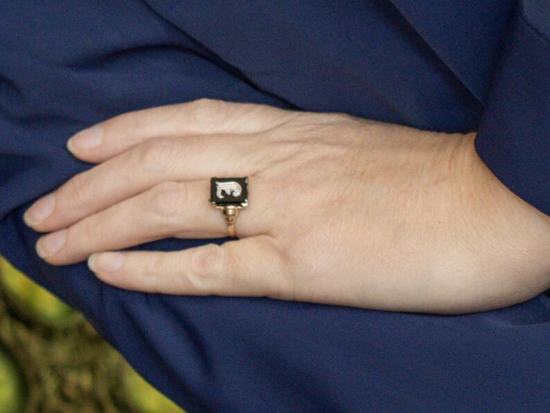 May include: A gold-toned ring with a black rectangular face featuring a white letter "E". The ring is set against a dark blue fabric background. The ring is a classic piece of jewelry.