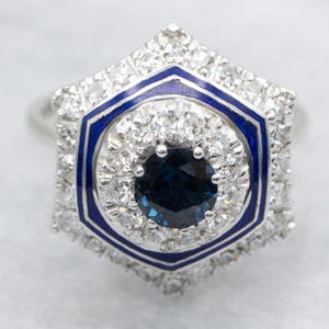 May include: A silver ring featuring a dark blue gemstone center, surrounded by a halo of smaller diamonds. The ring has a geometric design with a blue enamel border, creating a striking contrast.