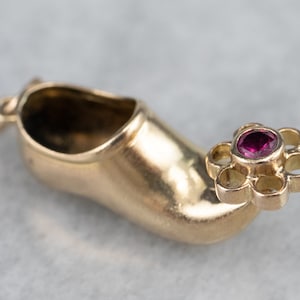 Puede incluir: Un amuleto de oro en forma de zapato con una flor de piedra preciosa roja en la punta.