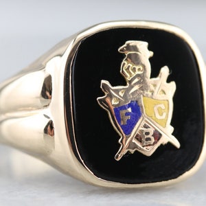 May include: A gold ring with a black onyx stone featuring a crest with the letters "F", "C", and "B" in gold and blue enamel.