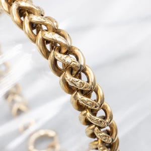 May include: Close-up of a gold-toned chain bracelet. The bracelet features a series of interlocking oval links, each adorned with delicate engraved patterns. The warm, metallic color of the chain contrasts against a soft, blurred background.