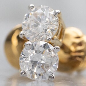 May include: A pair of stud earrings featuring round brilliant-cut clear stones set in four-prong white metal baskets. The earrings are secured with polished yellow gold screw-back posts, showcasing a classic design with high-clarity faceted stones.