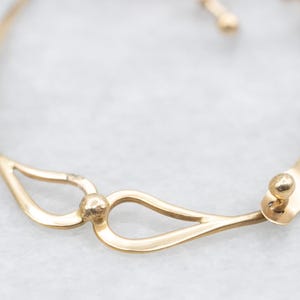 May include: A close-up view of a gold-colored bracelet. The bracelet features a delicate design with teardrop-shaped links and a central spherical accent. The metal has a polished finish, reflecting light.