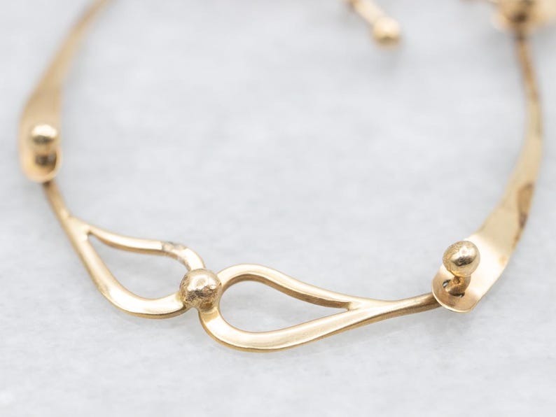May include: A close-up view of a gold-colored bracelet. The bracelet features a delicate design with teardrop-shaped links and a central spherical accent. The metal has a polished finish, reflecting light.