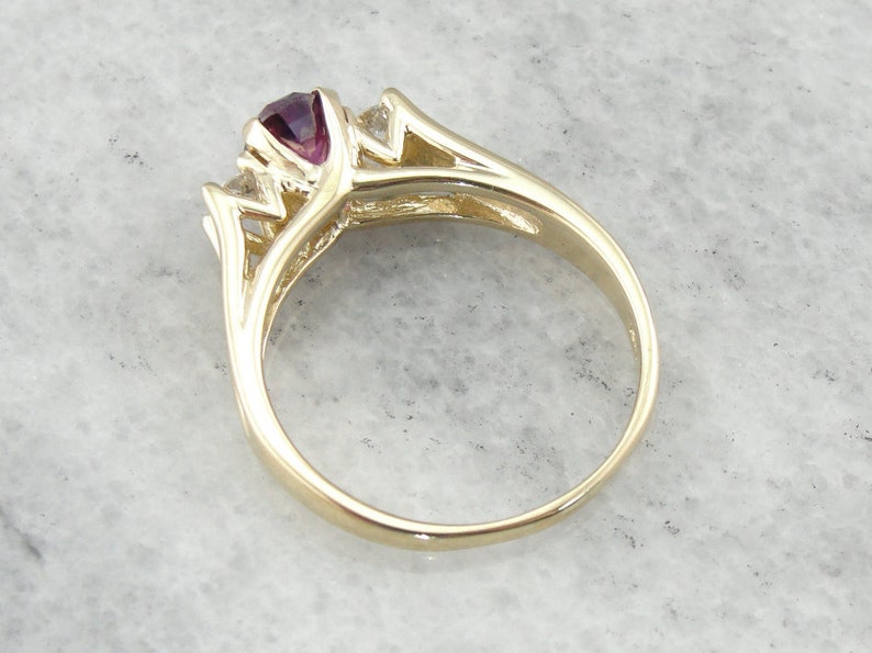 May include: A gold ring with a purple gemstone and two smaller clear gemstones. The ring has a delicate, intricate design.