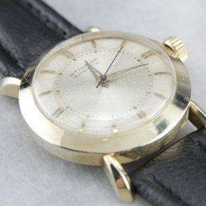 Pu&ograve; includere: Un orologio da polso dorato con quadrante bianco e cinturino in pelle nera. Il quadrante dell'orologio ha indici orari in numeri romani neri e la scritta "Wittnauer Automatic" stampata su di esso.