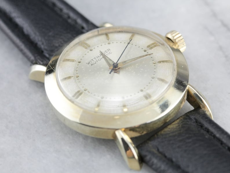 Pu&ograve; includere: Un orologio da polso dorato con quadrante bianco e cinturino in pelle nera. Il quadrante dell'orologio ha indici orari in numeri romani neri e la scritta "Wittnauer Automatic" stampata su di esso.
