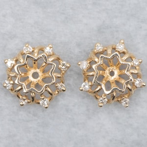 May include: Two gold-toned, star-shaped earrings with a floral design. Each earring features a central open circle surrounded by a star-shaped pattern and small, clear gemstones. The earrings are set against a light gray background.