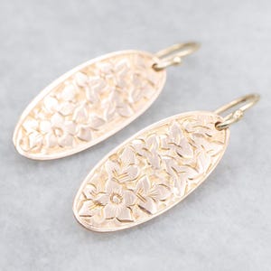 May include: A pair of gold-toned oval earrings. Each earring features a floral pattern across its surface. The earrings have a hook closure. The earrings are a light gold color.