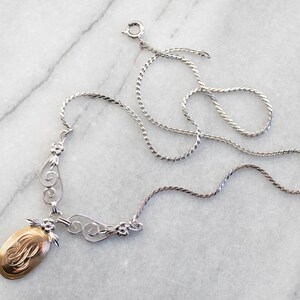 May include: A silver chain necklace with a gold oval pendant featuring a monogram. The pendant is attached to a delicate silver filigree design.