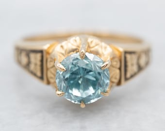 May include: A gold ring with a blue gemstone set in a detailed, ornate setting. The band is decorated with a black enamel design.