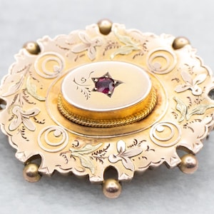 Puede incluir: Un broche dorado con una piedra roja en el centro. El broche está decorado con intrincados diseños florales y tiene un borde festoneado.