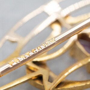 May include: Close-up of a gold-toned brooch with a geometric design. The brooch has a pin on the back and is marked "BRIAR" on the pin.