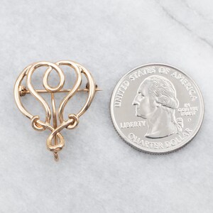 May include: A gold-toned, intricate, abstract design brooch. The brooch is shaped like a heart with a loop at the top. The brooch is about the size of a quarter.