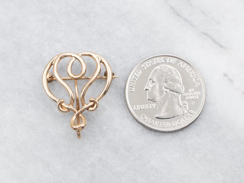 May include: A gold-toned, intricate, abstract design brooch. The brooch is shaped like a heart with a loop at the top. The brooch is about the size of a quarter.