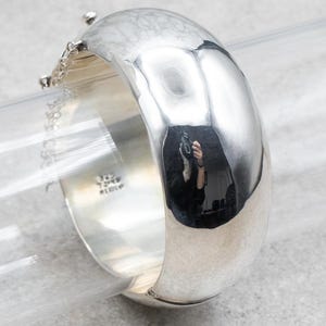 以下が含まれることがあります： 滑らかで磨かれた仕上げのシルバーのバングルブレスレット。ブレスレットは開いており、ヒンジ付きの留め具が付いています。