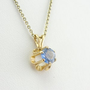 Op de afbeelding: Een gouden ketting met een hanger met een blauwe edelsteen in een gouden swirl-ontwerp.