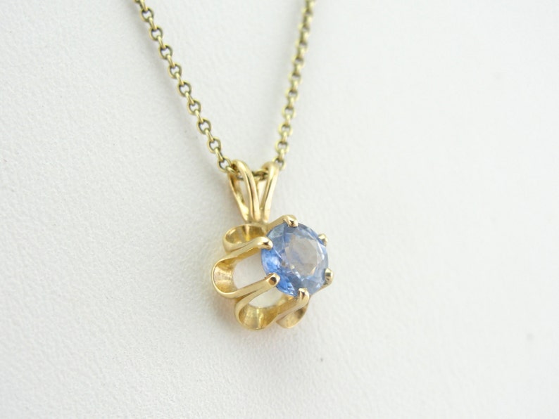 Op de afbeelding: Een gouden ketting met een hanger met een blauwe edelsteen in een gouden swirl-ontwerp.