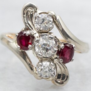 May include: A silver ring featuring three round stones. The central stone is a large diamond, with two smaller red ruby stones on either side.