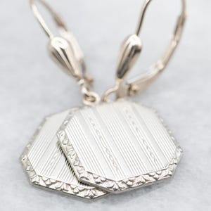 May include: A pair of silver octagon-shaped dangle earrings with a textured design. The earrings have a simple hook closure.