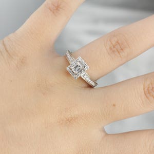 Puede incluir: Un anillo de plata con un diamante talla esmeralda de forma cuadrada rodeado de diamantes m&aacute;s peque&ntilde;os. El anillo tiene una banda que est&aacute; engastada con diamantes m&aacute;s peque&ntilde;os.