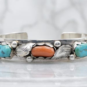 May include: A silver cuff bracelet featuring alternating oval turquoise and orange coral cabochons set in scalloped bezels. The band is decorated with polished silver leaf motifs and small silver spheres between each stone setting.