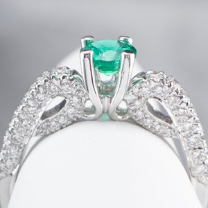 May include: A silver ring featuring a large emerald green gemstone surrounded by smaller diamonds. The ring has a delicate, intricate design.