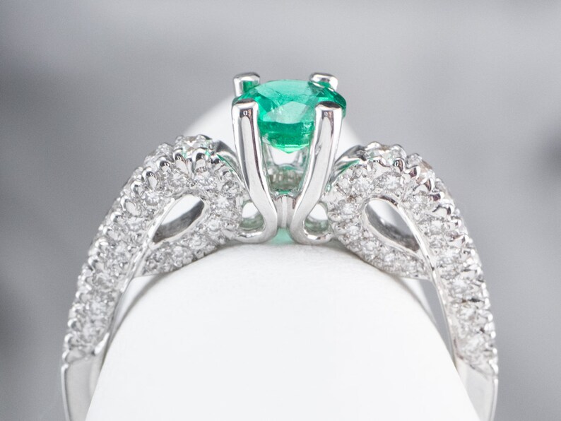 May include: A silver ring featuring a large emerald green gemstone surrounded by smaller diamonds. The ring has a delicate, intricate design.