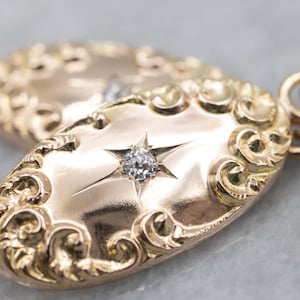 May include: Close-up of a gold-toned pendant with an oval shape. The pendant features a starburst design with a central diamond and ornate scrollwork around the edges. The background is a soft grey.
